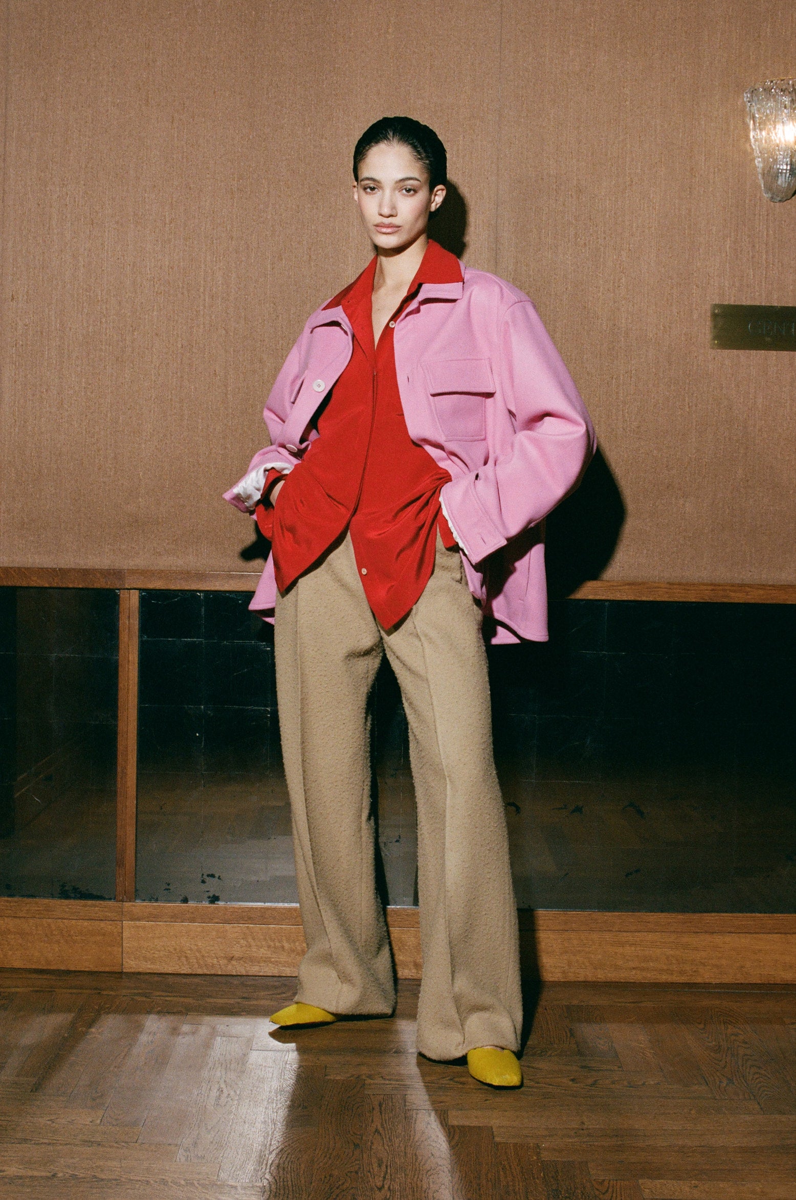 Woman wearing a pink jacket over a red shirt and beige pants in an indoor setting.