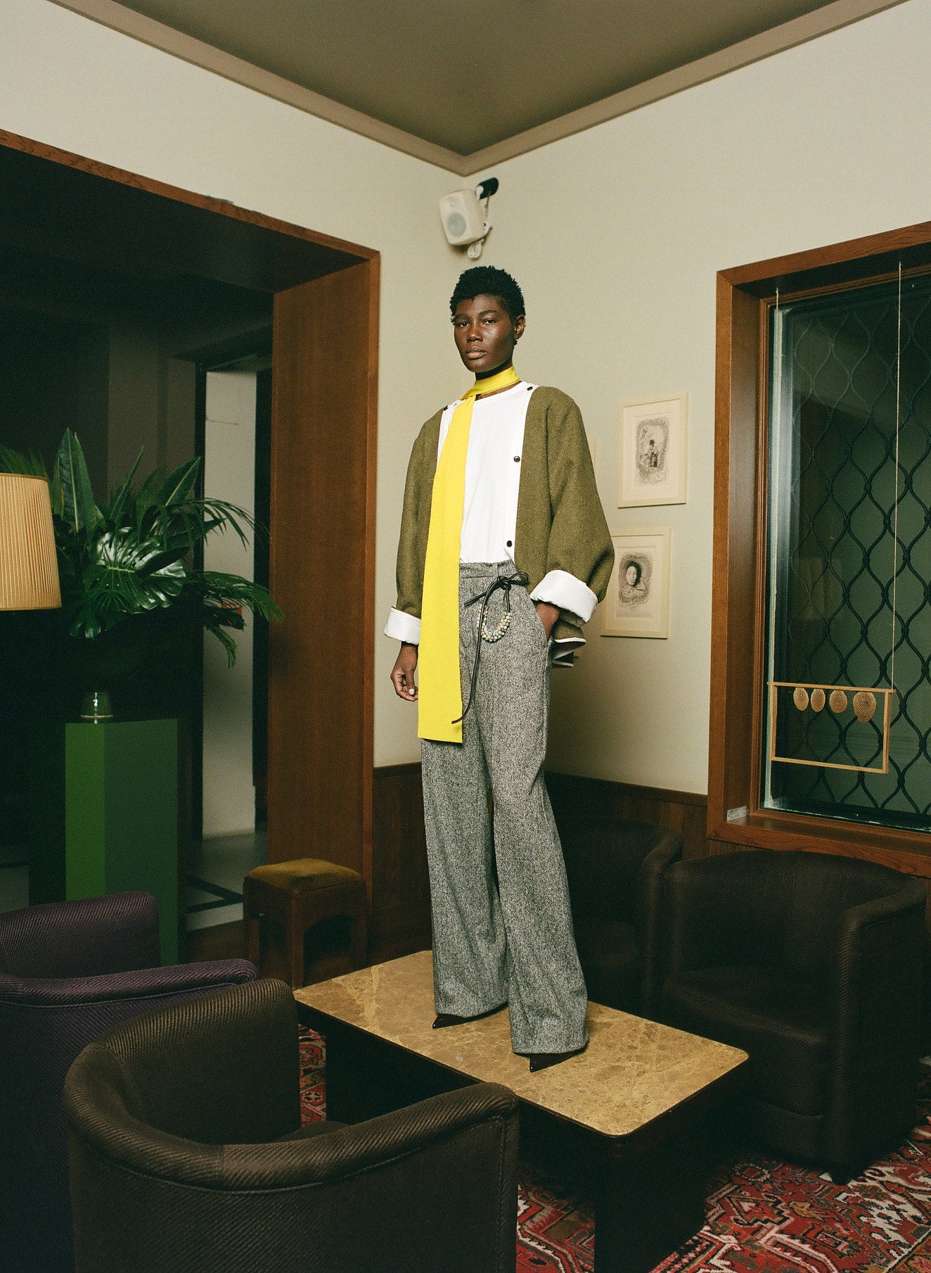 Person standing in a room with vintage decor, wearing a green cardigan, yellow shirt, and gray pants.