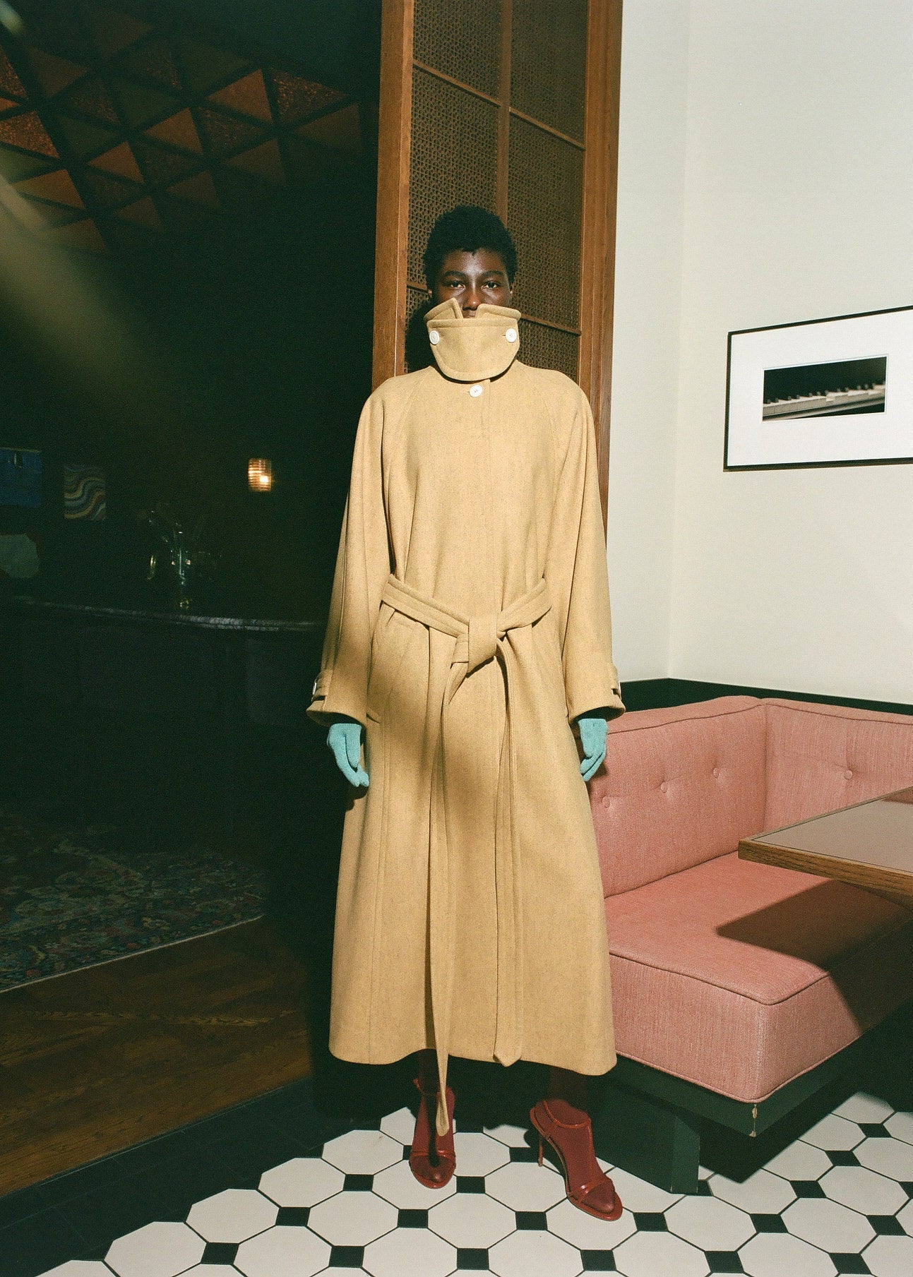 Person wearing a beige trench coat in an indoor setting with a pink couch and framed picture on the wall.