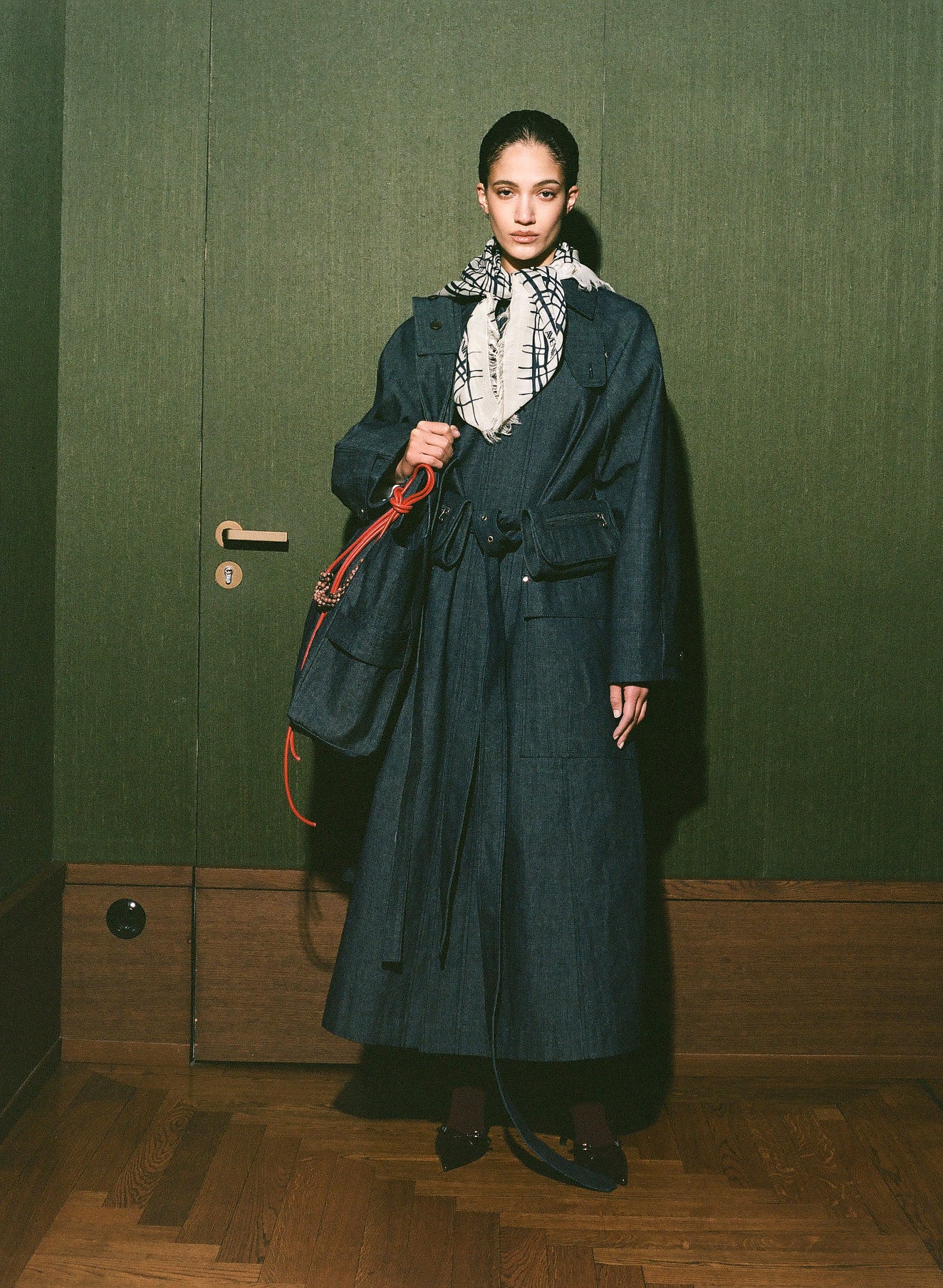 Woman in a long dark coat standing in a room with a green wall and wooden floor.